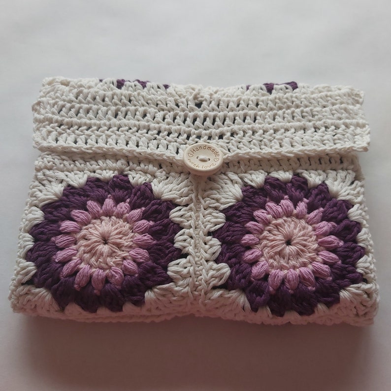 May include: A white crocheted pouch with a purple and pink floral design. The pouch has a button closure with the text "Handmade with Love".