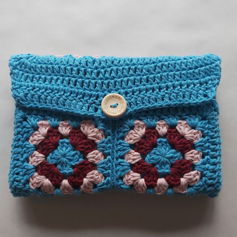 May include: A blue crocheted pouch with a pink and burgundy granny square pattern. The pouch has a wooden button with the text "Handmade with love" on it.