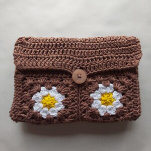 May include: A brown crocheted clutch with a flap closure and a wooden button. The clutch features two white crocheted daisies with yellow centers.