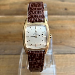 Könnte beinhalten: Eine goldfarbene Omega De Ville Automatik-Armbanduhr mit rechteckigem Zifferblatt und braunem Lederarmband. Das Zifferblatt ist silberfarben mit schwarzen Zeigern und Stundenmarkierungen. Das Armband hat ein strukturiertes Reptilienmuster.