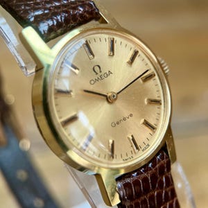 Pode incluir: Um relógio de pulso Omega Genève em tom dourado com um mostrador redondo cor de champanhe, marcadores de hora e ponteiros dourados. O relógio inclui uma pulseira de couro texturizado castanho escuro com um padrão de grão visível.