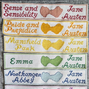 May include: Six white bookmarks with colorful embroidered bows and the titles of Jane Austen's novels: Sense and Sensibility, Pride and Prejudice, Mansfield Park, Emma, Northanger Abbey, and Persuasion.