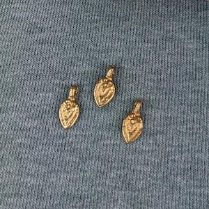 Peut inclure: Une boîte en bois avec un couvercle, remplie de petits charmes en métal de couleur dorée. Trois des charmes sont posés sur une surface en tissu bleu.