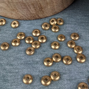 Può includere: Una ciotola di legno piena di piccole perline metalliche dorate e lucenti. Alcune perline sono sparse su una superficie di tessuto blu-grigio. La ciotola ha un coperchio con un design decorativo.