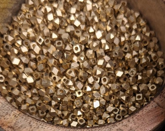 May include: A wooden bowl filled with gold-colored metal beads. The beads are small and have a pyramid shape. Some beads are scattered on a blue fabric surface.