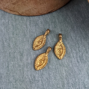 Puede incluir: Tres colgantes de color dorado con un diseño de ojo. Los colgantes están sobre una superficie de tela azul.