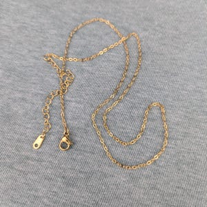 Peut inclure: Un collier doré délicat avec une fine chaîne et un fermoir mousqueton. Le collier est posé sur un tissu gris. La chaîne mesure environ 40 cm de long.