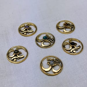Puede incluir: Una colección de seis colgantes dorados con el símbolo Om, cada uno con una piedra preciosa diferente en el centro. Las piedras preciosas incluyen púrpura, marrón, verde y gris. Los colgantes son circulares con un diseño ornamentado.
