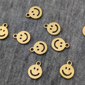 May include: Gold-colored smiley face charms with a small loop at the top for attaching to jewelry or other crafts. The charms have a simple, minimalist design with a curved smile and two dots for eyes. The charms are scattered across a gray surface.