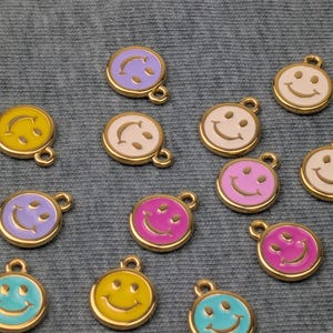 Peut inclure: Une collection de breloques smiley colorées avec des bords dorés. Les breloques présentent différentes couleurs, dont le jaune, le rose, le violet et le turquoise, chacune avec un motif de visage souriant. Ces breloques sont probablement utilisées pour la fabrication de bijoux ou l'artisanat.
