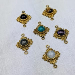 Puede incluir: Seis abalorios de metal dorado con diferentes cabujones de piedras preciosas. Los abalorios tienen forma cuadrada con detalles ornamentados y tres lazos para sujetarlos a joyas.