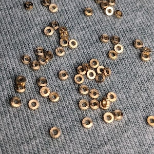 Può includere: Primo piano di una superficie di tessuto grigio con molti piccoli anelli di metallo dorato sparsi su di essa.