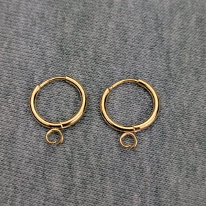 May include: A pair of gold-colored hoop earrings. Each earring has a circular hoop with a small, attached loop at the bottom. The earrings are displayed on a textured, gray surface.