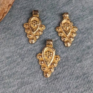 May include: Three gold-coloured metal pendants with a detailed, ornate design. Each pendant features a teardrop shape with a textured surface and a small loop at the top for attaching to a chain or cord.