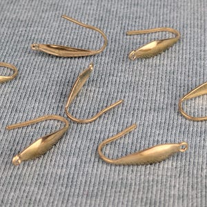 Pode incluir: Close-up de vários ganchos de brincos dourados. Os ganchos têm um design curvo com uma base em forma de gota e um pequeno laço na extremidade. O fundo é um tecido azul claro texturizado.