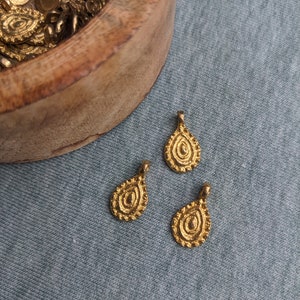 May include: Three gold-colored teardrop-shaped pendants with intricate designs. The pendants are on a blue fabric background.