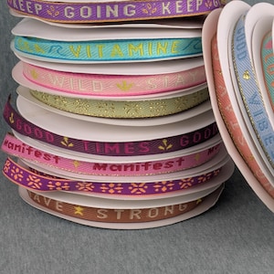 Könnte beinhalten: Ein Stapel dekorativer Bandspulen in verschiedenen Farben, darunter Lila, Türkis, Rosa und Braun. Jedes Band zeigt inspirierende Texte wie "Keep Going" und "Good Vibes". Geeignet für Bastelarbeiten und Geschenkverpackungen.