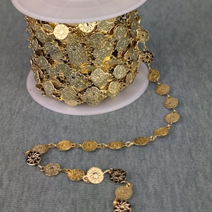 Könnte beinhalten: Eine Spule mit goldfarbener Kette mit einem sich wiederholenden Sonnenmuster. Die Kette besteht aus kleinen, flachen, kreisförmigen Gliedern. Die Kette ist teilweise abgewickelt und zeigt das detaillierte Muster.
