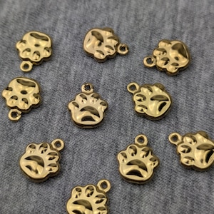Op de afbeelding: Goudkleurige pootafdrukbedels met een lus voor het maken van sieraden. De bedels zijn klein, met gedetailleerde afdrukken van voetzolen en klauwen. De bedels zijn verspreid over een grijze ondergrond.