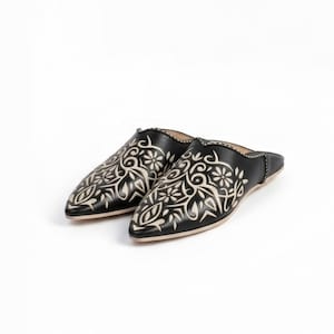 May include: A pair of black leather slippers with a pointed toe and a cream-colored floral design. The slip-on shoes are decorated with an intricate, stylized botanical pattern.