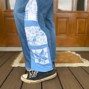 May include: Blue denim jeans featuring a vertical side panel constructed from a patchwork of light blue and white fabrics. The panel includes patterns of small bows, floral motifs, gingham checks, and a central star design in blue and white.