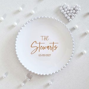 May include: A round white ceramic plate with a beaded edge. The center features the brown text THE Stewarts and the date 03/05/2027. The plate is surrounded by scattered white spherical beads arranged in a heart shape and individual loose beads.