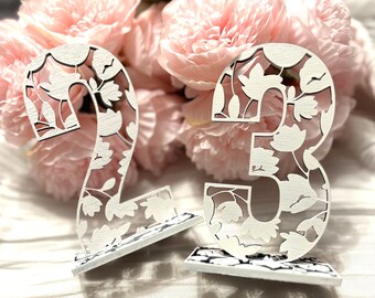 May include: Two white wooden table numbers, 2 and 3, with a floral design. The numbers are on white stands.