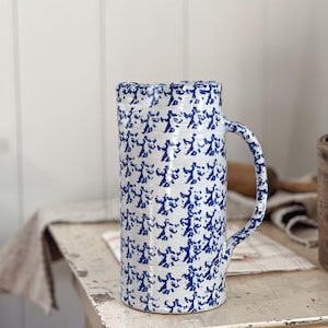 Puede incluir: Una jarra de cerámica blanca con un patrón floral azul y blanco. La jarra tiene una boca ancha y un asa.