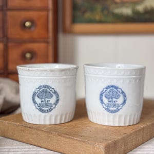 May include: Two white ceramic containers with a blue tree logo and text. The containers have a ridged base and a decorative rim. The logo reads "MILANDES PASTES FRAMERIES M C". They are placed on a wooden cutting board.
