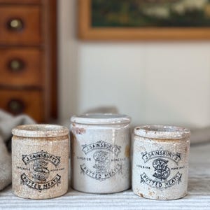May include: Three cylindrical ceramic pots with a cream-colored crackle glaze finish. Each pot features black printed text reading J. SAINSBURY'S SUPERIOR POTTED MEATS with a central letter S logo. The pots have a uniform shape and a vintage appearance.