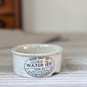 Può includere: Un piccolo vasetto in ceramica color bianco sporco di forma cilindrica con base piatta. L'esterno presenta un'etichetta ovale stampata in nero con la scritta MADE ONLY BY W.A.TURNER BOND ST. STOWMARKET, con decorazioni sopra e sotto il testo.