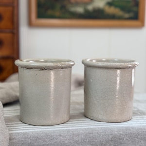 May include: Two cylindrical ceramic crocks with a light gray, glazed finish. Each vessel features a thick, rounded rim at the top and a smooth, straight-sided body. The surface of the right crock displays a visible network of fine crackle patterns.