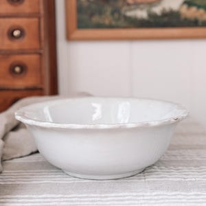Puede incluir: Un cuenco blanco de estilo antiguo con borde festoneado. El cuenco tiene una superficie ligeramente texturizada y está sobre una tela a rayas. El fondo incluye un mueble de madera y un cuadro enmarcado, sugiriendo una estética vintage.