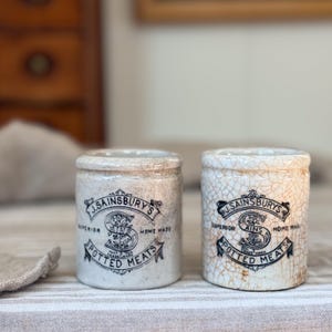 May include: Two cylindrical ceramic pots with a cream-colored crackle glaze finish. Each pot features black printed text reading J. SAINSBURY'S SUPERIOR HOME MADE POTTED MEATS with a central letter S and floral details within a decorative banner design.