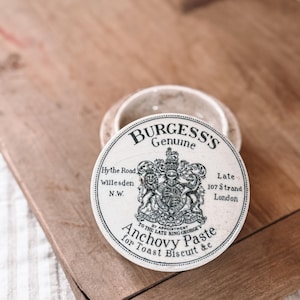 May include: A vintage, round, white container lid with the text "BURGESS'S Genuine Anchovy Paste for Toast Biscuit &c." The lid features a detailed crest and text indicating its origin in London. The container sits on a wooden surface.
