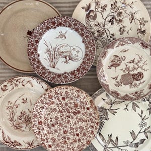 May include: A collection of antique ceramic plates with various brown and white floral and scenic designs. The plates feature intricate patterns, including flowers, birds, and oriental-inspired motifs. The plates are arranged on a striped surface.