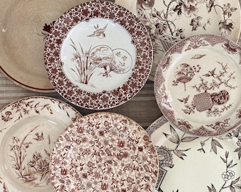 May include: A collection of antique ceramic plates with various brown and white floral and scenic designs. The plates feature intricate patterns, including flowers, birds, and oriental-inspired motifs. The plates are arranged on a striped surface.