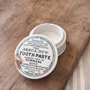 May include: An antique white ceramic Areca Nut Tooth Paste container with the lid slightly ajar. The lid features black text that reads "Areca Nut Tooth Paste" and other text. The container sits on a wooden surface.