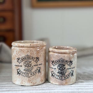 May include: Two cylindrical ceramic pots with a cream-colored crackle glaze finish. Each pot features black printed text reading J. SAINSBURY'S SUPERIOR HOME MADE POTTED MEATS with a central letter S logo. The pots vary in size, standing side by side.