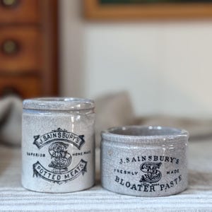 May include: Two cylindrical ceramic pots with a light grey crackle glaze finish. The taller pot features black text reading J. SAINSBURY'S SUPERIOR HOME MADE POTTED MEATS. The shorter pot features black text reading J. SAINSBURY'S FRESHLY MADE BLOATER PASTE.