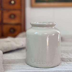 May include: A cylindrical, light gray ceramic jar with a smooth, glossy finish and a slightly flared rim. The base of the jar features the embossed text "LAB-Lagny" in capital and lowercase letters. The vessel has a wide opening and a sturdy base.