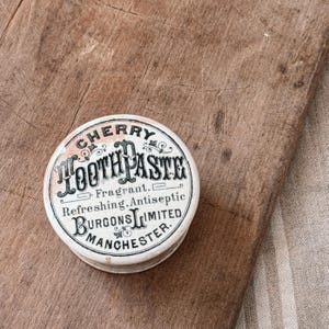 May include: A vintage, round, white and cream-colored tin of "Cherry Toothpaste" with black and red lettering. The tin reads "Fragrant, Refreshing, Antiseptic" and "Burgons Limited, Manchester."