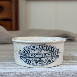 May include: A vintage off-white ceramic pot with a black printed label that reads G.W. PLUMTREE MANUFACTURER OF HOME POTTED MEATS 13 RAILWAY ST SOUTHPORT. The cylindrical container features a slightly flared rim and a weathered, aged appearance.