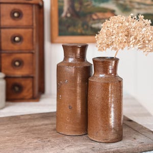 Puede incluir: Dos jarrones de gres marrón con un aspecto rústico y envejecido. Un jarrón es más alto que el otro. Un arreglo floral seco está en el jarrón más pequeño. Los jarrones están sobre una superficie de madera, con un mueble de madera y una obra de arte enmarcada en el fondo.