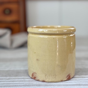May include: A small, cylindrical, antique stoneware jar with a light yellow glaze. The jar has a rounded rim and a slightly textured surface. The base of the jar has a few reddish-brown spots. The background is a striped fabric.