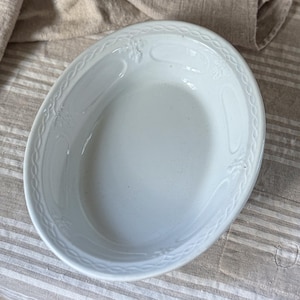 May include: Oval white serving dish with a decorative border and embossed details. The dish has a smooth, glossy surface and is set against a neutral striped background. A classic piece for serving food.