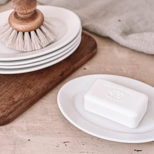 May include: A white oval soap dish with a white bar of soap inside. The soap has the text "LA TOILETTE" embossed on it. The dish is sitting on a light pink surface.