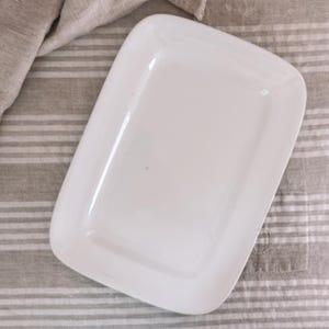May include: A rectangular white ceramic serving platter with rounded corners and a smooth, glossy finish. The platter features a flat, recessed center area and a wide, slightly raised rim, providing a clean and simple design for food presentation.