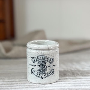 May include: A small, cylindrical white ceramic pot with a distinct crackle glaze finish. The front features black printed text that reads J. SAINSBURY'S SUPERIOR HOMEMADE POTTED MEATS, centered around a large decorative letter S with floral details.