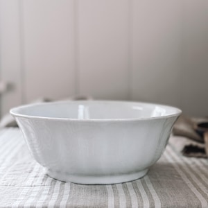 May include: A white ceramic bowl with a wide, shallow design. The bowl has a slightly flared rim and sits on a small, rounded base. The bowl is on a striped fabric.